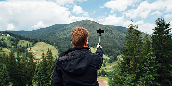 Junger Mann macht Bild von sich selbst. Im Hintergrund sind ein bewaldeter Berg und Wiesen zu sehen.