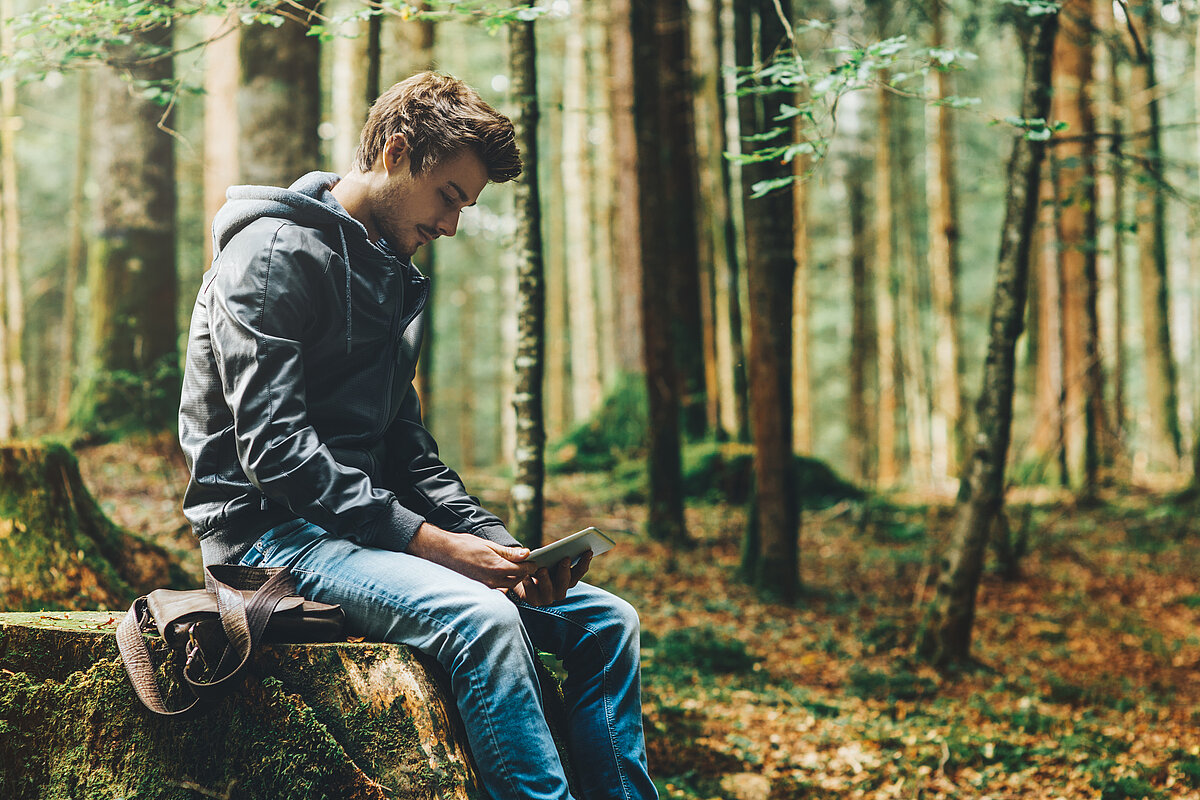 Ein junger Mann sitzt im Wald und benutzt ein digitales Tablet