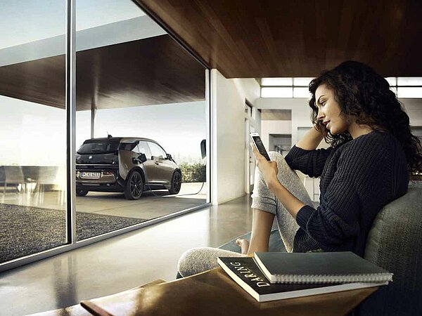 Frau sitzt im Wohnzimmer und steuert mit ihrem Telefon die Ladung ihres Elektroautos. Im Hintergrund ist das Elektroauto, das an der Ladestation angeschlossen ist, zu sehen.