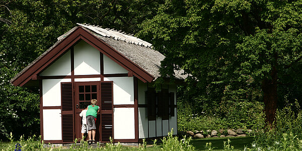 Zwei Kinder schauen in kleines Häuschen in der Natur zum Fenster hinein.