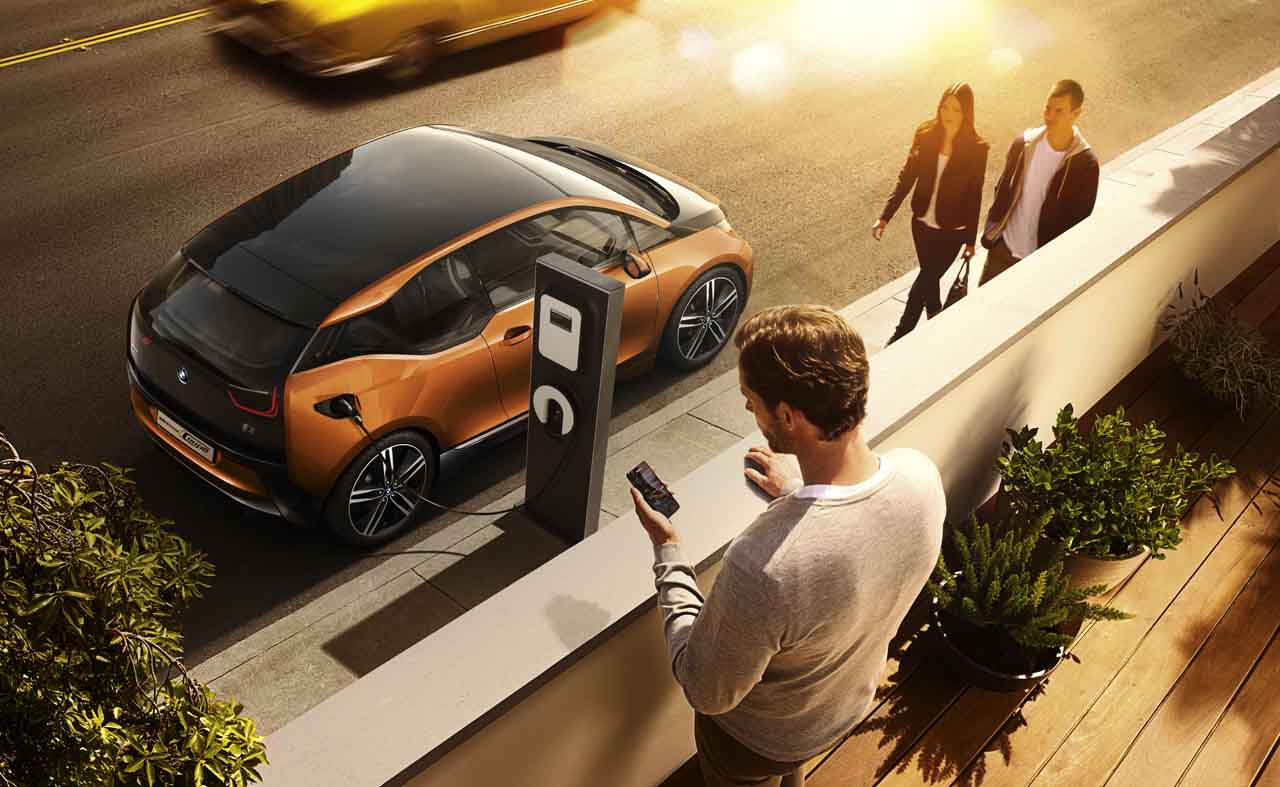 Mann steht auf Balkon und steuert mit seinem Telefon die Ladung seines Elektroautos. Im Hintergrund sind das Elektroauto angeschlossen an der Ladestation auf der Straße und ein spazierendes Paar zu sehen.